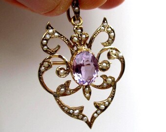Puede incluir: Un colgante de oro con una amatista de color violeta claro de corte ovalado en el centro, rodeada por un intrincado marco calado decorado con numerosas pequeñas perlas blancas. La pieza incluye una anilla en la parte superior.