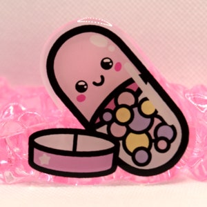 May include: A pink pill shaped charm with a smiling face and colorful dots inside. The pill is open and a small pink pill container with a star is next to it.