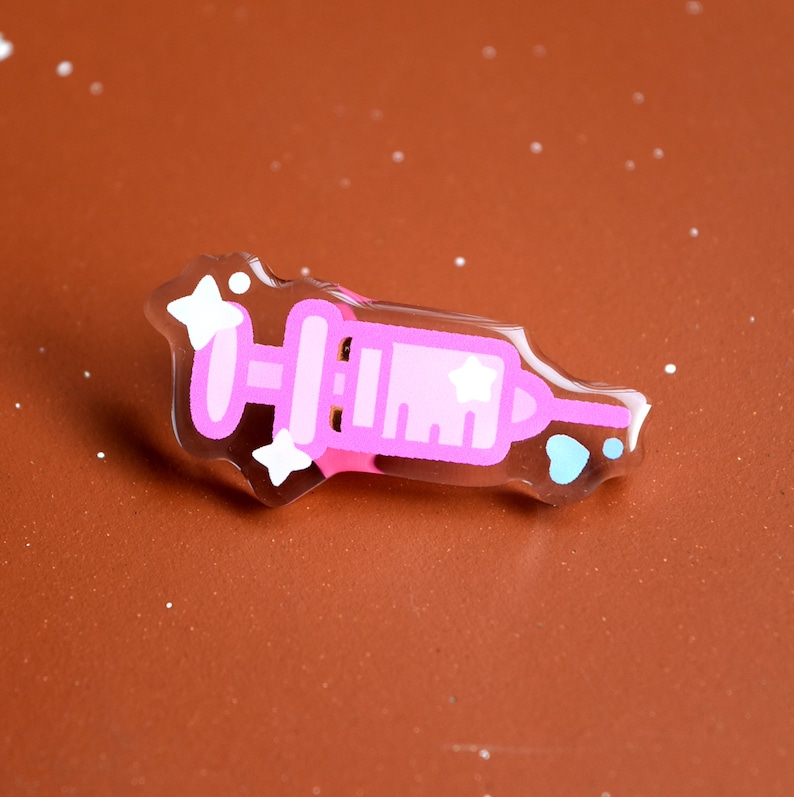 May include: A pink and white syringe with a star on the side, surrounded by white stars, in a clear plastic frame.