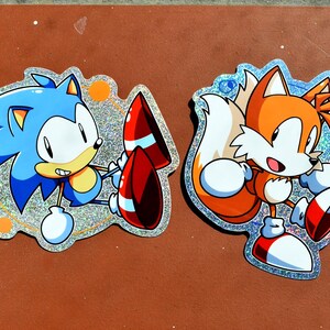 May include: Two stickers featuring Sonic the Hedgehog and Tails from the Sonic video game series. Sonic is blue with a red and white rocket. Tails is orange and white. Both stickers have a glittery, iridescent border.