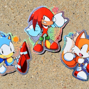 May include: Three holographic stickers featuring cartoon characters from the Sonic the Hedgehog series. One sticker shows a blue hedgehog sitting, another shows a red echidna standing with fists raised, and the third shows an orange fox running.