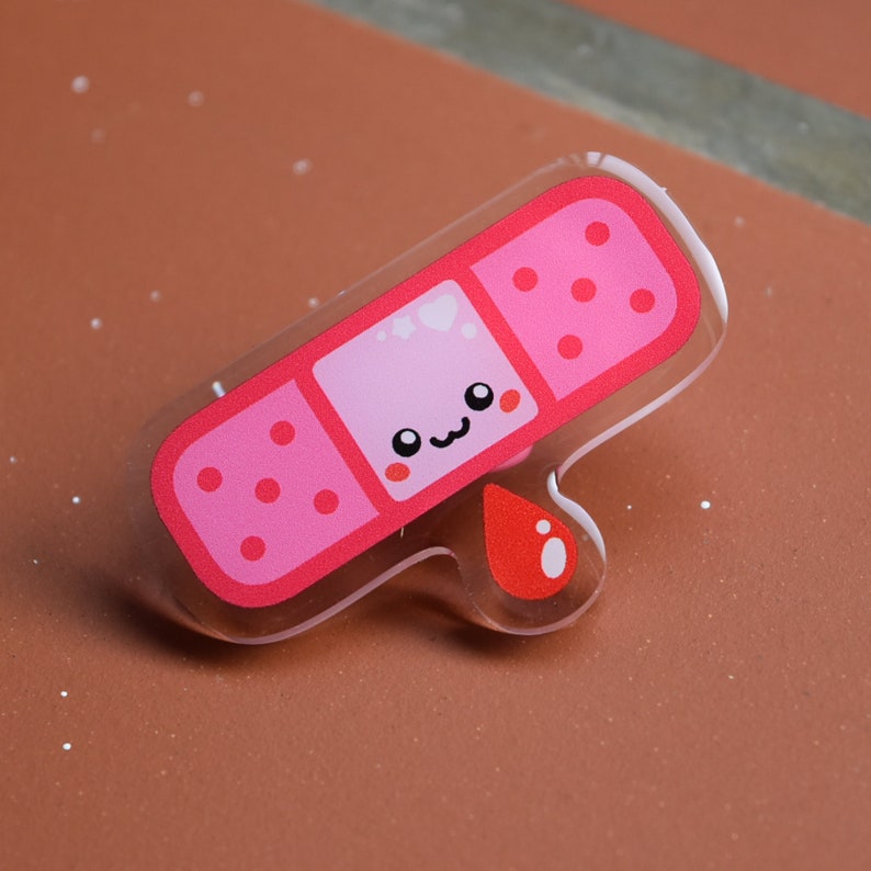 May include: A pink and red cartoon bandaid with a smiling face and a red drop of blood. The bandaid is made of clear acrylic.