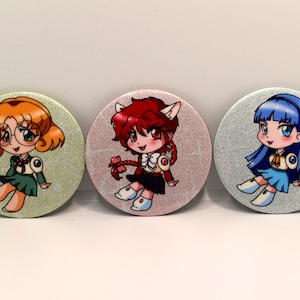 May include: Three round pinback buttons with glitter backgrounds featuring cartoon anime girls. The first button has a green background with a girl with orange hair wearing a green dress and glasses. The second button has a pink background with a girl with red hair wearing a white and red dress. The third button has a blue background with a girl with blue hair wearing a blue dress.