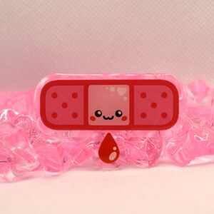 May include: A pink and red cartoon bandaid with a cute face and a red blood drop. The bandaid is on a pink background.