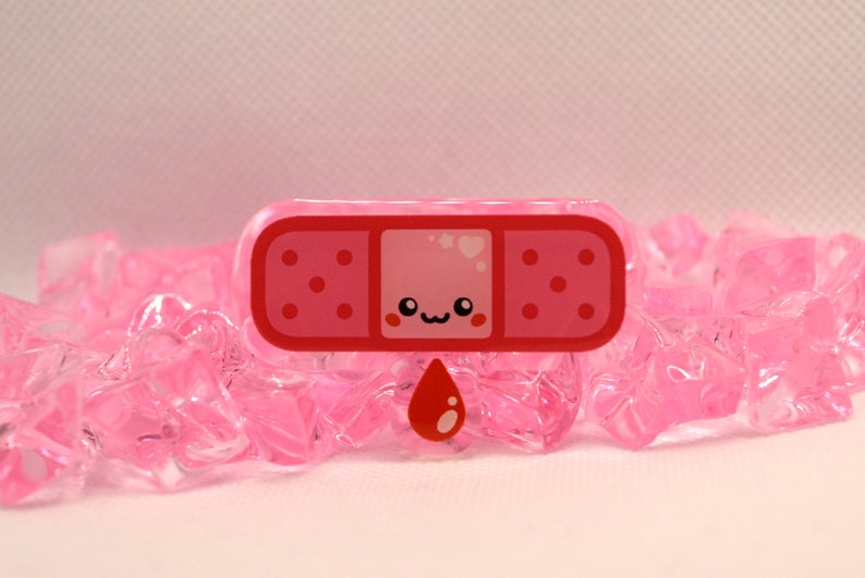 May include: A pink and red cartoon bandaid with a cute face and a red blood drop. The bandaid is on a pink background.