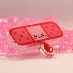 May include: A pink and red cartoon band-aid with a cute face and a single red blood drop. The band-aid is made of clear acrylic.