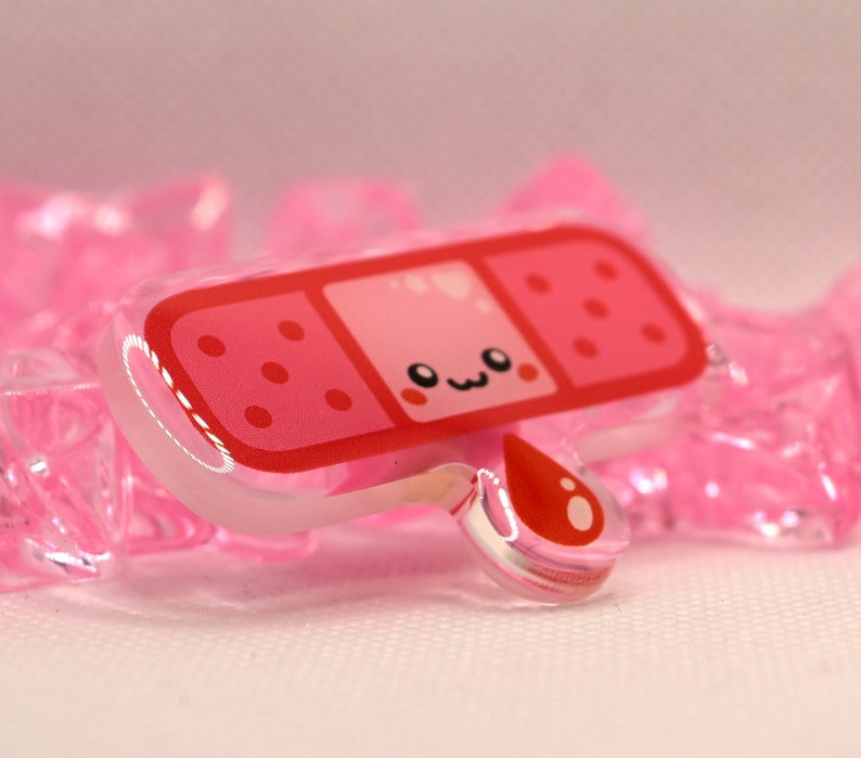 May include: A pink and red cartoon band-aid with a cute face and a single red blood drop. The band-aid is made of clear acrylic.