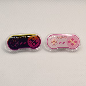 Puede incluir: Dos pines de plástico transparente con forma de mando de videojuego. El pin de la izquierda tiene un mando negro con un degradado rosa y amarillo. El pin de la derecha tiene un mando rosa sobre fondo blanco. El pin de la izquierda tiene el texto "コントローラー" en japonés. El pin de la derecha tiene el texto "かわいい" en japonés.
