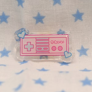 May include: A pink and white plastic pin with a retro video game controller design. The pin has two blue hearts on either side of the controller.
