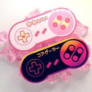 Könnte beinhalten: Zwei Aufkleber in Form von Videospiel-Controllern. Der obere Aufkleber ist hellrosa mit gelben Tasten und dem japanischen Text "かわいい". Der untere Aufkleber zeigt einen Farbverlauf von Dunkelblau zu Pink mit schwarzen Tasten und dem Text "コアゲーマー".