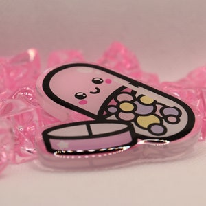 May include: A pink and white pill shaped charm with a smiling face and colorful candies inside. The charm is made of clear acrylic and has a glossy finish.
