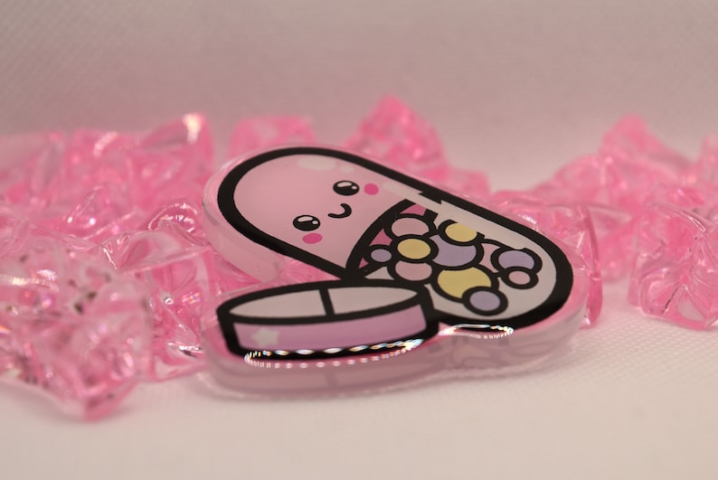 May include: A pink and white pill shaped charm with a smiling face and colorful candies inside. The charm is made of clear acrylic and has a glossy finish.