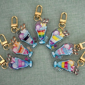 Może przedstawiać: Dziewięć kolorowych breloków do kluczy ze złotymi kółkami. Każdy brelok ma przezroczysty plastikowy zawieszkę z kreskówkową ilustracją mlecznego koktajlu z różnymi kolorowymi warstwami i dodatkami.