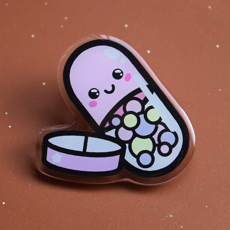 May include: A pink pill shaped charm with a smiling face and colorful dots inside. The pill is open and the bottom half is visible.
