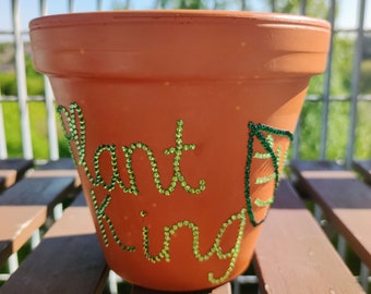 May include: A terracotta flower pot with the words "Plant King" spelled out in green rhinestones. A green rhinestone leaf design is on the right side of the pot.