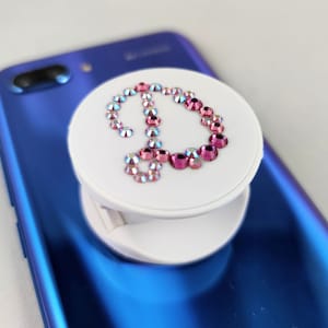May include: A white phone grip with a pink and iridescent crystal design in the shape of a peace sign.