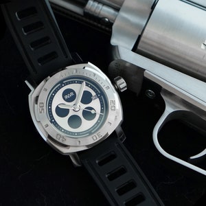 Op de afbeelding: Een zilveren revolver met een zwarte greep en een zilveren horloge met een zwarte band en een witte wijzerplaat met zwarte markeringen. De wijzerplaat van het horloge heeft het woord "RLVR" erop.