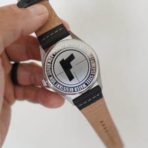 May include: Back of a silver watch with a black leather band. The watch face has the text "WATER RESISTANT 30M JAPAN MOV RECON SNIDER SERIES STAINLESS STEEL" around a target with a black "F" in the center.