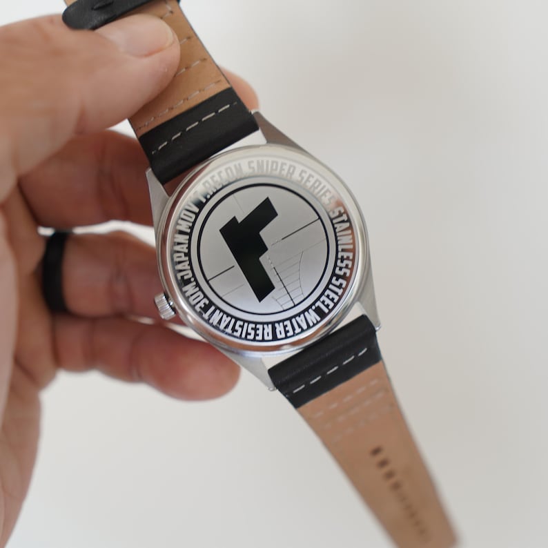 May include: Back of a silver watch with a black leather band. The watch face has the text "WATER RESISTANT 30M JAPAN MOV RECON SNIDER SERIES STAINLESS STEEL" around a target with a black "F" in the center.