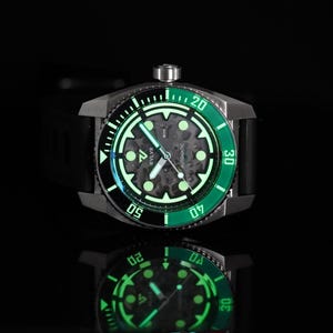 Op de afbeelding: Een zwart en zilveren automatisch horloge met een groene bezel en een skeletwijzerplaat. Het horloge is verlicht met een groen materiaal dat in het donker gloeit.