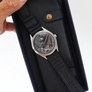 May include: A black leather watch band with a silver watch face featuring a map design. The watch is in a black box with a black leather wallet.