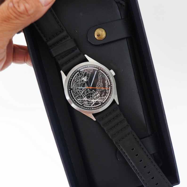 May include: A black leather watch band with a silver watch face featuring a map design. The watch is in a black box with a black leather wallet.