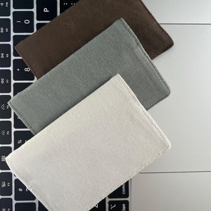 Puede incluir: Tres artículos rectangulares de tela en tonos marrón, gris y blanquecino están dispuestos sobre un teclado de portátil. Los artículos parecen ser pequeñas carteras o tarjeteros, con una superficie texturizada y bordes cosidos.