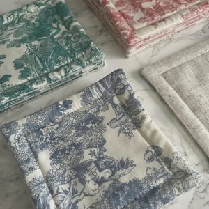 May include: A collection of square fabric coasters featuring toile de jouy patterns in teal, red, and blue on white backgrounds, alongside a single plain beige linen coaster. Each coaster has a thick, quilted border and a soft, layered construction.