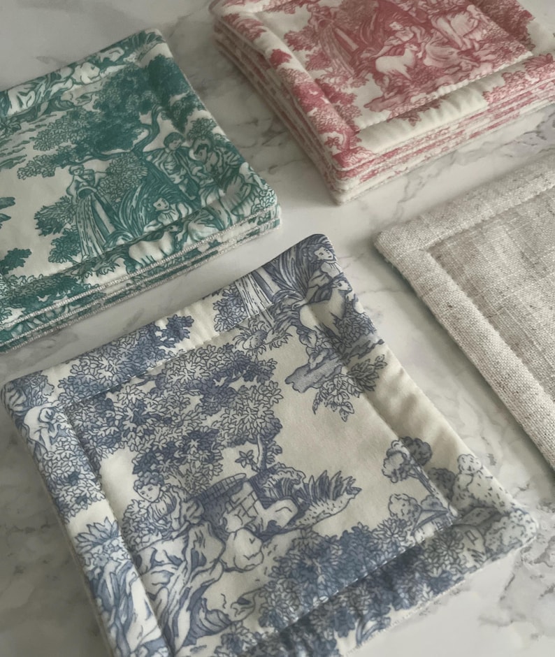 May include: A collection of square fabric coasters featuring toile de jouy patterns in teal, red, and blue on white backgrounds, alongside a single plain beige linen coaster. Each coaster has a thick, quilted border and a soft, layered construction.