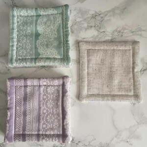 May include: Three square fabric coasters with stitched borders. One coaster features a light green floral lace pattern, another displays a lavender floral lace design, and the third is a solid, textured beige linen-like fabric with visible weave.