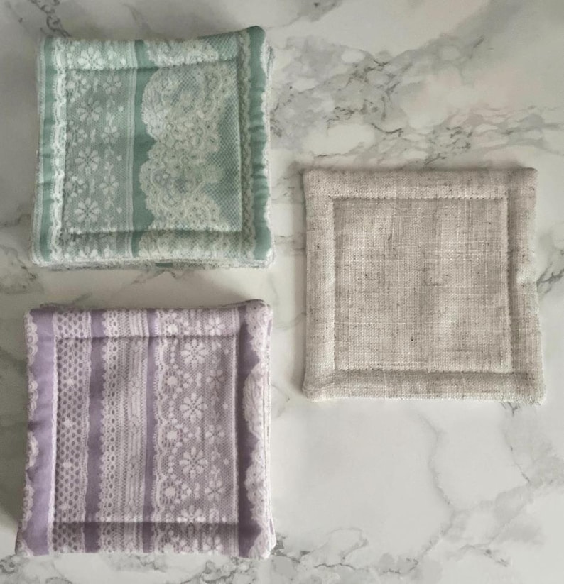 May include: Three square fabric coasters with stitched borders. One coaster features a light green floral lace pattern, another displays a lavender floral lace design, and the third is a solid, textured beige linen-like fabric with visible weave.