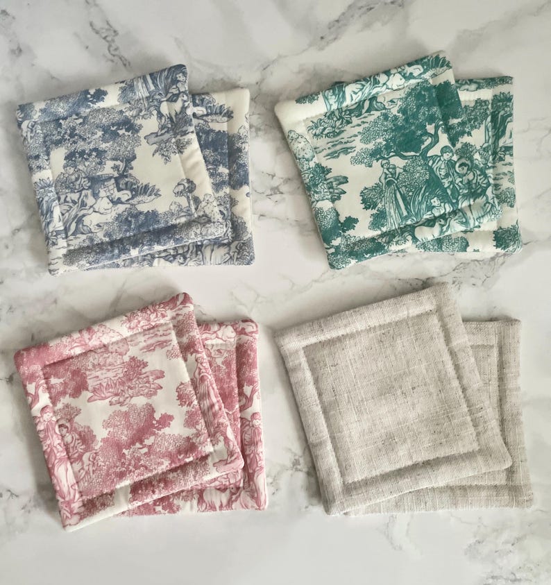 May include: A set of eight square fabric coasters arranged in four pairs. Three pairs feature a toile de jouy pattern in blue, green, and pink on a cream background. The fourth pair is made of a plain, textured, light beige natural linen fabric.