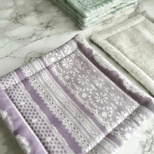 May include: A set of square fabric coasters featuring a lavender and white lace-patterned design, a solid beige linen-textured coaster, and a stack of light green floral-patterned coasters, all with stitched borders and a quilted construction.