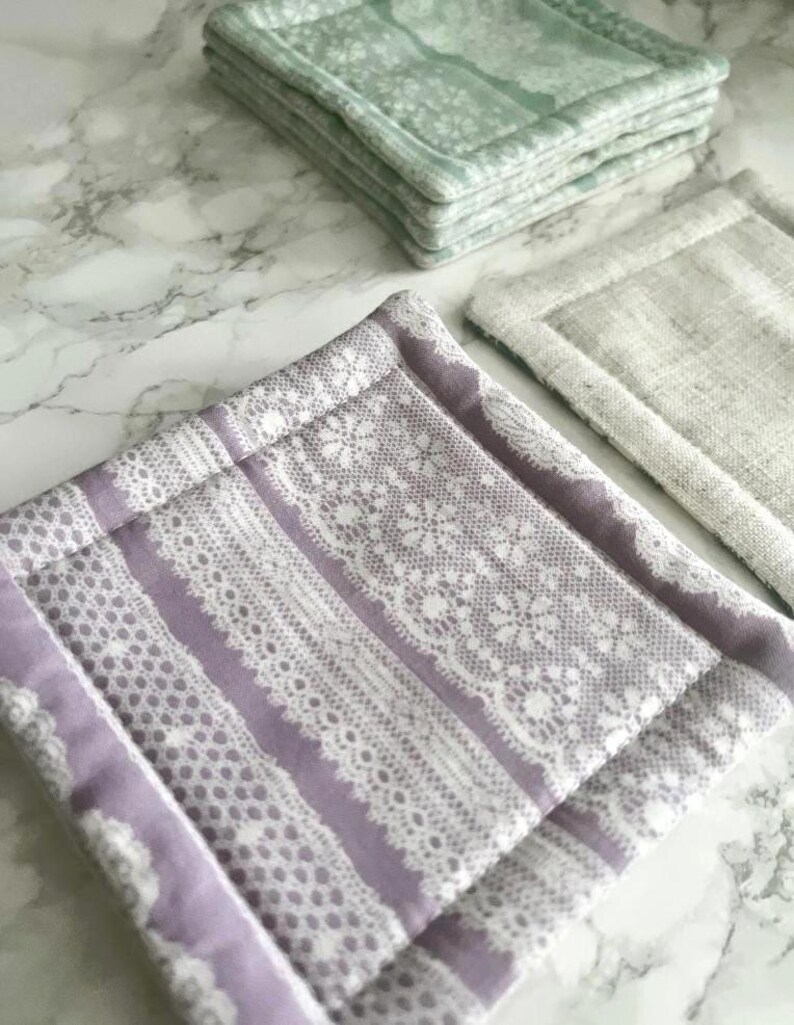 May include: A set of square fabric coasters featuring a lavender and white lace-patterned design, a solid beige linen-textured coaster, and a stack of light green floral-patterned coasters, all with stitched borders and a quilted construction.