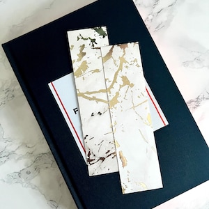 May include: A black book with a white and gold marble design bookmark and a white and red striped label. The items are on a white and gray marble surface.
