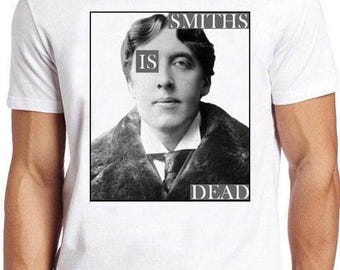 May include: A white short-sleeved t-shirt featuring a black and white portrait of Oscar Wilde. The graphic includes the text "SMITHS" at the top right, "IS" over the left eye, and "DEAD" at the bottom right, all enclosed within a black rectangular border.