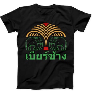 May include: A black short-sleeve t-shirt featuring a graphic print of two green elephants facing each other under a stylized yellow palm tree. A small red brick-like icon sits between the elephants, with green Thai text printed below the design.