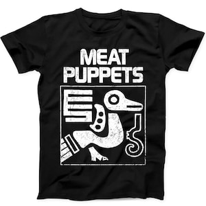 May include: A black short-sleeve t-shirt featuring the white text MEAT PUPPETS above a square graphic. The graphic contains a stylized white bird-like figure with geometric patterns and a distressed, weathered texture throughout the print.
