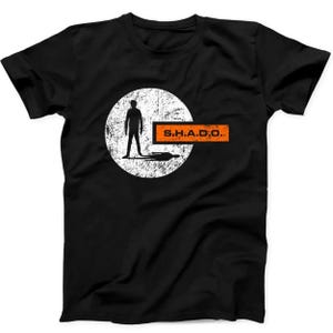 May include: A black short-sleeved t-shirt featuring a graphic design with a white distressed circle containing a black silhouette of a person standing. An orange rectangular bar extends from the circle with the black text S.H.A.D.O. printed inside.
