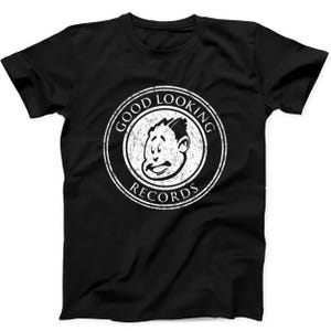 Puede incluir: Una camiseta negra de manga corta con un gráfico circular blanco en el pecho. El diseño incluye una cara de dibujos animados estilizada en el centro, rodeada por el texto GOOD LOOKING RECORDS en una fuente desgastada y envejecida.