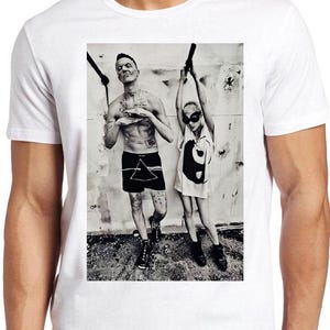 Puede incluir: Una camiseta blanca de manga corta con un estampado fotográfico en blanco y negro en la parte delantera. El gráfico muestra a dos personas de pie contra una pared texturizada, una con pantalones cortos oscuros y la otra con una prenda clara.