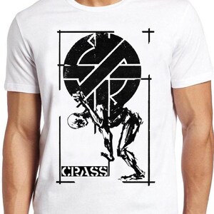 Peut inclure: Un t-shirt blanc à manches courtes avec une impression graphique noire. Le motif représente une silhouette stylisée soutenant un grand emblème circulaire avec une croix. Le texte CRASS est imprimé dans un rectangle en bas du motif.