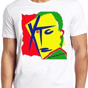 May include: A white short-sleeved t-shirt featuring a graphic print of a stylized yellow face with blue facial features, a green textured hair section, and a red background. The text XTC is printed in blue, and the text drums and wires is in black.