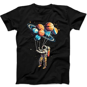 Puede incluir: Una camiseta negra de manga corta con un estampado gráfico de un astronauta con traje blanco flotando en el espacio. El astronauta sostiene cuerdas unidas a un grupo de planetas coloridos que parecen globos sobre un fondo estrellado.
