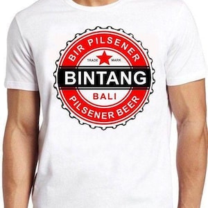 May include: A white short-sleeve t-shirt featuring a circular red and black graphic on the chest. The design resembles a bottle cap and includes the text BIR PILSENER, TRADE MARK, BINTANG, BALI, and PILSENER BEER with a central red star.