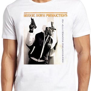 Może przedstawiać: Biały t-shirt z krótkim rękawem z nadrukiem graficznym okładki albumu By All Means Necessary zespołu Boogie Down Productions. Nadruk przedstawia osobę w kurtce i czapce trzymającą broń palną, z napisem BOOGIE DOWN PRODUCTIONS oraz BY ALL MEANS NECESSARY.