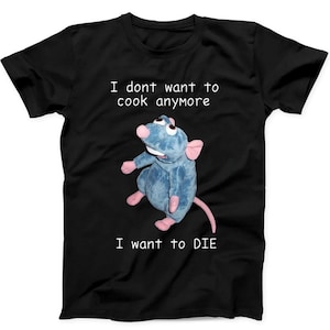 Puede incluir: Una camiseta negra de manga corta con un personaje de rata de peluche azul en el centro. El texto sobre la rata dice "I dont want to cook anymore" y el texto debajo de la rata dice "I want to DIE" en una fuente blanca sin remates.