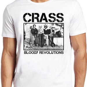 May include: A white short-sleeved t-shirt featuring a black and white rectangular graphic print on the chest. The graphic displays a group of people standing outdoors with the text CRASS at the top and BLOODY REVOLUTIONS at the bottom.