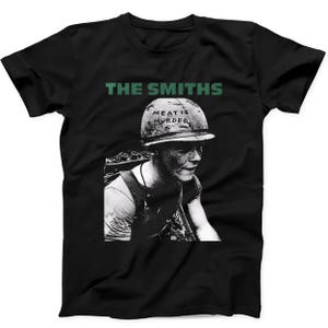 May include: A black short-sleeve t-shirt featuring a green text graphic that reads THE SMITHS above a black and white illustration of a soldier wearing a helmet with the text MEAT IS MURDER written on the front of the helmet surface.