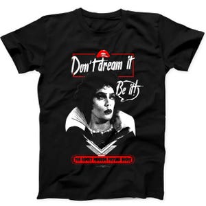 May include: A black short-sleeved t-shirt featuring a graphic of Dr. Frank-N-Furter from The Rocky Horror Picture Show. The print includes the text Don't dream it, Be it! in white, a red pair of lips, and the title in a red rectangular frame.